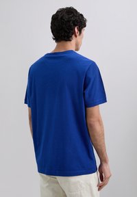 Hombre con cabello oscuro rizado y corto, vistiendo una camisa de manga corta azul lisa y pantalones crema, de pie de espaldas contra un fondo claro y liso.