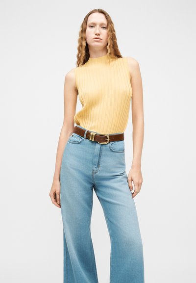 Femme aux longs cheveux ondulés portant un haut sans manches jaune côtelé, un jean large taille haute bleu clair, et une ceinture marron avec une boucle dorée.