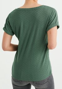 Grön texturerad t-shirt med fiskbensmönster, korta uppvikta ärmar och avslappnad passform, sedd bakifrån.