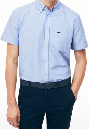 Homme portant une chemise bleu clair à manches courtes avec un bouton-pression et une poche poitrine, ainsi qu'un pantalon bleu foncé avec une ceinture texturée.