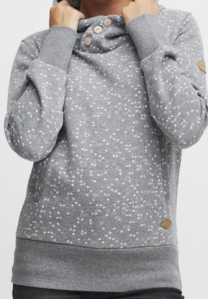 Personne portant un pull gris avec des petits motifs en points multicolores, ajustant le col haut avec les mains près du cou.
