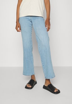 Person trägt hellblaue, gerade geschnittene Jeans, schwarze offene Sandalen mit Zehenfreiheiten und ein weißes Oberteil, steht vor einem schlichten hellen Hintergrund.