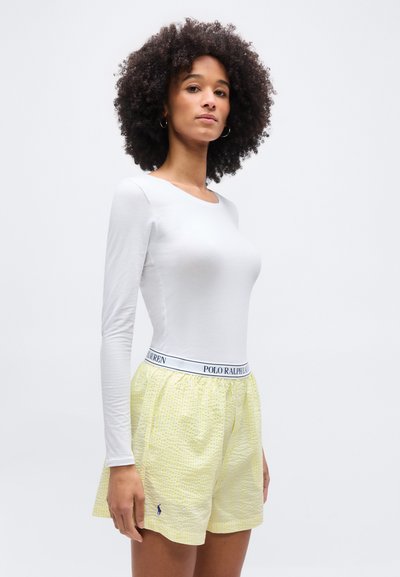 Γυναίκα με σγουρά μαλλιά που φοράει λευκό μακρυμάνικο τοπ και κίτρινο σορτς με υφή και μέση Polo Ralph Lauren.