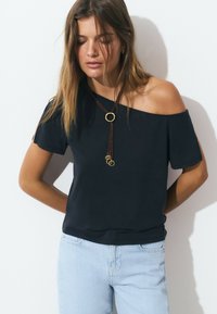 Haut noir à épaules dénudées en tissu doux, avec des manches courtes, un pendentif circulaire accentué en or et une coupe décontractée. Associé à un jean en denim clair.