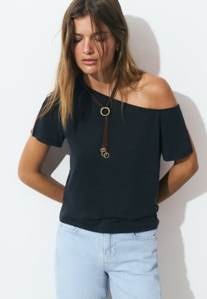 Top negro de hombros al aire hecho de una tela suave, con mangas cortas, un colgante en forma de círculo dorado y un corte relajado. Combinado con jeans de mezclilla claros.