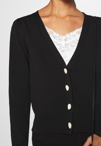 Cardigan noir en tricot avec un col en V, doté de quatre boutons de fermeture dans un matériau contrastant, et d'un ourlet côtelé. Superposé à un haut en dentelle blanche.