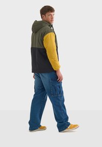 Chaqueta con bloques de color que presenta secciones en verde oliva, amarillo y negro. Fabricada con un material ligero, cuenta con una capucha y se combina con pantalones cargo azules y zapatillas amarillas.