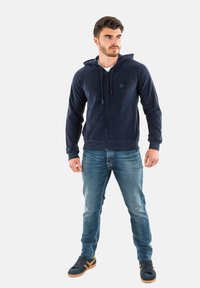Marineblauwe zip-up hoodie van zacht materiaal met een trekkoordhoed, voorzakken en logo-detail. G gedragen met lichtblauwe jeans en donkere sneakers.