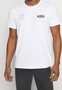White cotton t-shirt featuring a round neckline, short sleeves, and black printed branding on the chest and sleeve.