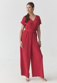 Tuta rossa strutturata con scollo a V, maniche corte e vita con cintura; design a gambe larghe; accessoriata con un bracciale dorato e sandali.