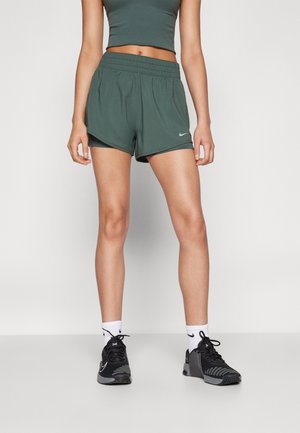 Frau trägt dunkelgrüne Sportshorts und passendes Crop-Top, weiße Kniestrümpfe und schwarze Laufschuhe, steht vor einfarbigem Hintergrund.