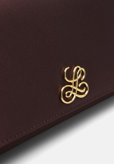 Logo monogramme entrelacé doré brillant sur une surface en cuir lisse marron foncé avec des coutures visibles le long du bord.