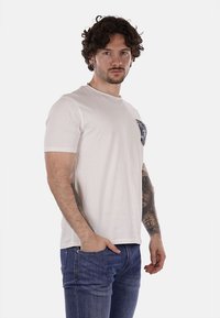 Camiseta blanca de manga corta con un cuello redondo, que presenta un bolsillo estampado en el lado izquierdo. La tela parece suave; el modelo lleva puestos unos pantalones vaqueros azules.