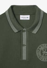 Ciemnozielona koszulka polo z przeszywanym kołnierzykiem i białymi paskami, z haftowanym logo na klatce piersiowej oraz zapięciem na dwa guziki.