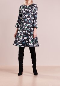 Abito a maniche svasate nero con stampa floreale, caratterizzato da fiori bianchi e dettagli verdi. Indossato con stivali al ginocchio in suede nero.