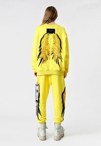 Sudadera amarilla y pantalones a juego con diseños de alas en negro y dorado. Presenta una etiqueta en el costado y una textura suave. Calzado blanco.