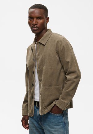 Homme portant une veste en velours côtelé marron clair par-dessus une chemise blanche et un jean bleu, une main dans la poche, sur un fond uni.