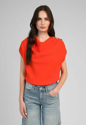 Femme aux longs cheveux bruns portant un haut sans manches rouge vif avec des boutons aux épaules et un jean taille haute bleu clair, debout devant un fond uni.