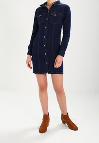 Kvinna som bär en åtsittande mörkblå denimklänning med knappar och långa ärmar, samt bruna ankle boots.