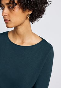 Ciemnoniebieska bluzka z długim rękawem, okrągłym dekoltem i gładką teksturą, w zestawie z dużymi złotymi kolczykami w kształcie koła. Skupienie na ramieniu i dekolcie.