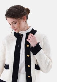 Elfenbeinfarbener Tweed-Jackett mit marineblauen Akzenten, runder Kragen und goldenen Knöpfen. Verfügt über Fransenverzierungen an den Manschetten und Taschen für zusätzlichen Struktur.