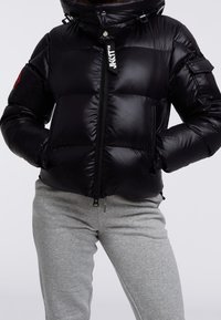 Czarna kurtka puchowa z błyszczącym wykończeniem, zapięciem na zamek, dwoma kieszeniami po bokach, odpinanym kapturem oraz czerwoną naszywką z logo na rękawie.