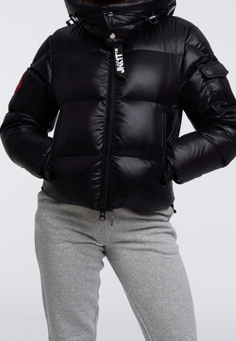 Czarna kurtka puchowa z błyszczącym wykończeniem, zapięciem na zamek, dwoma kieszeniami po bokach, odpinanym kapturem oraz czerwoną naszywką z logo na rękawie.