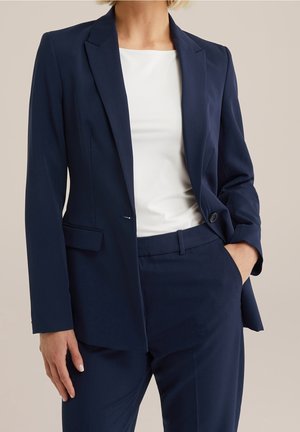 Femme portant un blazer bleu marine et un pantalon assorti avec une blouse blanche, debout avec une main dans la poche sur un fond uni.
