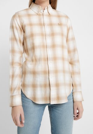 Kobieta ubrana w beżowo-białą koszulę w kratę zapinaną na guziki z kołnierzykiem oraz jasnoniebieskie dżinsy, stojąca na jednolitym tle.