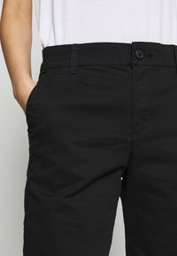 Personne portant un pantalon noir avec un bouton et une fermeture éclair, main gauche dans la poche, et une chemise blanche rentrée visible à la taille.