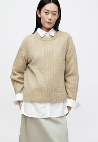 Μπεζ πλεκτό πουλόβερ με ριμπ υφή, φαρδιά μανίκια και στρογγυλή λαιμόκοψη, φορεμένο πάνω από ένα λευκό πουκάμισο με γιακά.