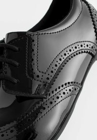 Schwarzer Lacklederschuh mit filigranen Brogue-Perforationen, flachen schwarzen Schnürsenkeln und einer glatten, glänzenden Oberfläche.