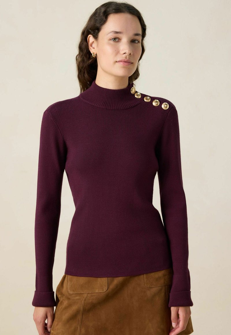 Femme portant un pull ajusté en ribbed prune avec des boutons dorés sur l'épaule gauche et une jupe en daim marron, se tenant devant un fond beige.