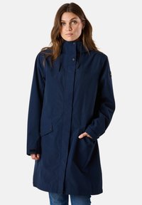 Woman wearing a navy blue long raincoat with high collar, snap buttons, flap pockets, and one hand in pocket, standing against a plain background.