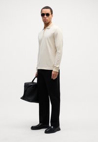 Beige langermet polo med krage, svarte bukser, svarte lærskjønner og en svart teksturert veske. Modell med mørke solbriller.
