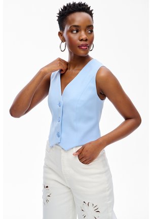 Kobieta o krótkich włosach, nosząca kolczyki koła, bezrękawnik w jasnoniebieskim kolorze z guzikami oraz białe spodnie z kwiatowymi wycięciami.