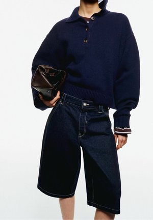 Person iført navyblå kort sweater og mørk denim knælange shorts, der holder en sammenfoldet brun lædertaske i hånden.