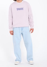 Felpa lilla chiaro con testo grafico blu, abbinata a jeans azzurri a vestibilità rilassata. Il modello indossa sneakers bianche. Stile semplice e casual.