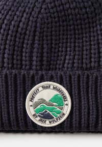 Beanie lavorato a maglia di un blu navy scuro con una texture a costine, caratterizzato da una toppa circolare di tessuto con motivi di montagne e animali in verde e grigio.