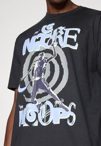 Μαύρο μπλουζάκι με γραφιστικό σχέδιο που απεικονίζει μια χορευτική φιγούρα και στροβιλιζόμενο φόντο. Η εκτύπωση περιλαμβά νει τις λέξεις "NIKE" και "WOOPS" σε μπλε τόνους.