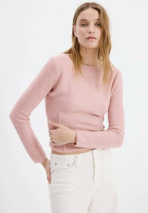 Pull rose tricoté à manches longues avec un col rond et des détails de couture sur les côtés, associé à un jean blanc. Texture douce et coupe ajustée.