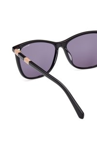 Schwarze Sonnenbrille im Cat-Eye-Design mit lila Gläsern und goldenen Akzenten an den Scharnieren. Glatter Kunststoffrahmen und schlanke schwarze Bügel.