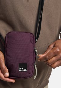 Paarse nylon schoudertas met rits, verstelbare strap en Jack Wolfskin-logo. Gladde textuur, compact rechthoekig ontwerp.