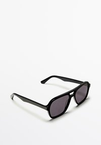 Schwarze rechteckige Sonnenbrille mit dunkel getönten Gläsern, die auf einer weißen Oberfläche liegt.