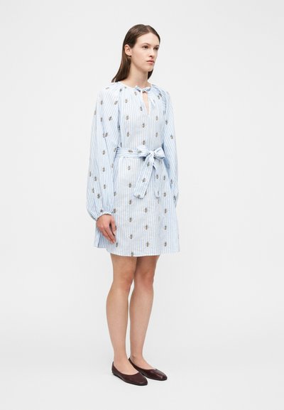 Γυναίκα όρθια φοράει ένα φόρεμα με λευκές και ανοιχτό μπλε ρίγες, τυπωμένες μέλισσες, μακριά μανίκια, ζώνη στη μέση που δένει και σκούρα επίπεδα παπούτσια σε λευκό φόντο.