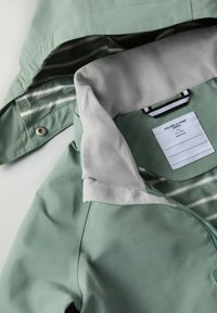 Vihreä takki, jossa on pehmeä harmaa kaulus ja verkkomateriaali vuorina. Sisältää brändin tunnuksen, vetoketjun ja nappikiinnityksen.