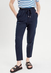 Pantalons en lin légers, bleu marine, avec taille à cordon, poches latérales et jambes fuselées, associés à des sandales noires à boucle.