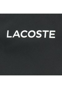 Zwarte stof met opvallende witte tekst "LACOSTE". Het materiaal heeft een gestructureerde afwerking, wat de duurzaamheid en stijl benadrukt.