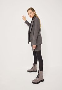 Mujer con blazer y falda gris, medias negras y botas de cordones gris claro, de pie contra una pared blanca con una mano tocándola.