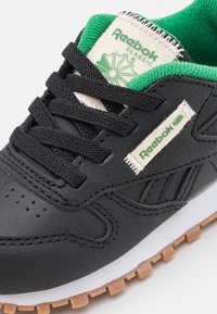 Svart lädersneaker med gröna accenter, texturerad yta, snörning och en vit mellansula. Har Reebok-logotypen broderad på sidan.